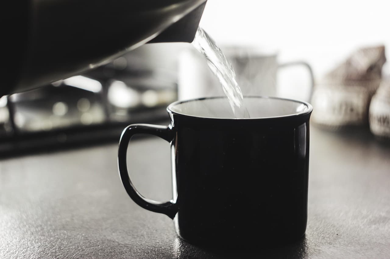 Hot water poured from a kettle into a black mug on a kitchen worktop